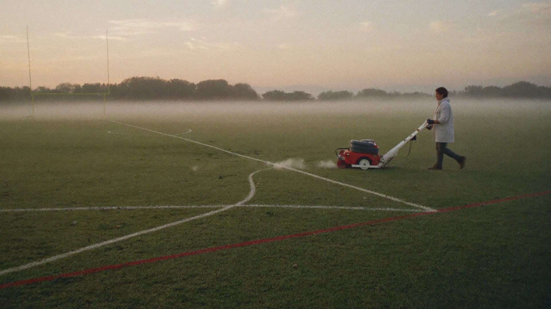 A groundskeeper chalking a fresh touchline across a misty pitch at dawn.